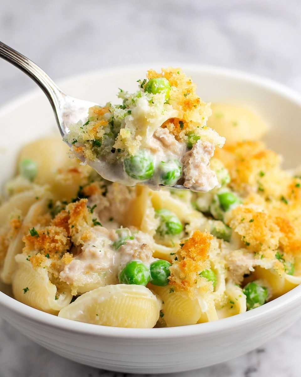 This image shows a close-up of a white bowl filled with a creamy pasta dish made with shell-shaped pasta. The pasta is covered with a thick white sauce and mixed with bright green peas and small pieces of light beige meat. On top, there is a golden-brown crunchy topping that looks like breadcrumbs with a crumbly texture. A silver spoon is holding some of the pasta, sauce, peas, meat, and crispy topping above the bowl. The bowl is set on a white marbled surface. photo taken with an iphone --ar 4:5 --v 7