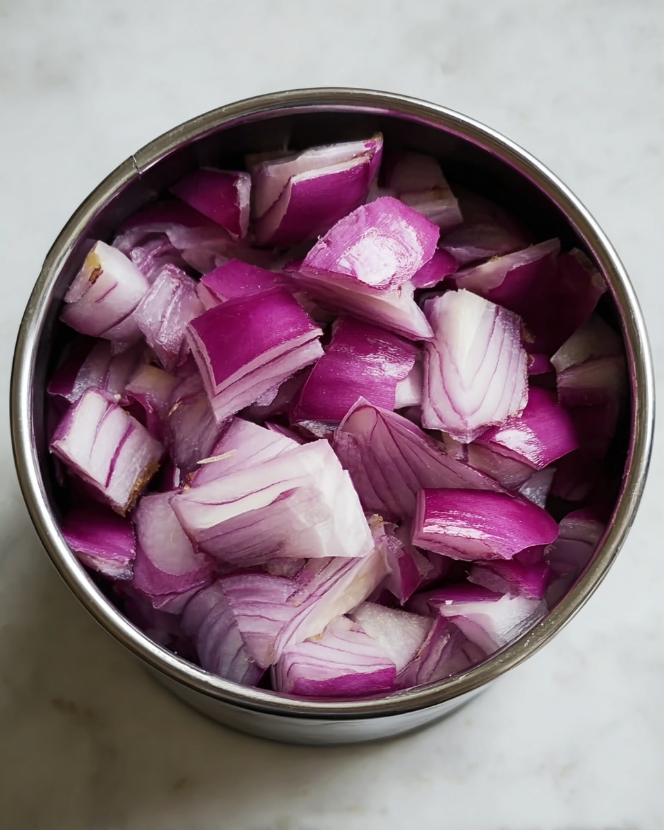 A close-up view of a metal container filled with chopped pieces of raw red onion, showing layers of purple and white with a shiny, smooth texture. The pieces are uneven in size and shape, filling the container to the top. The container is placed on a white marbled surface, adding a clean, bright contrast to the deep purple of the onion pieces. photo taken with an iphone --ar 4:5 --v 7