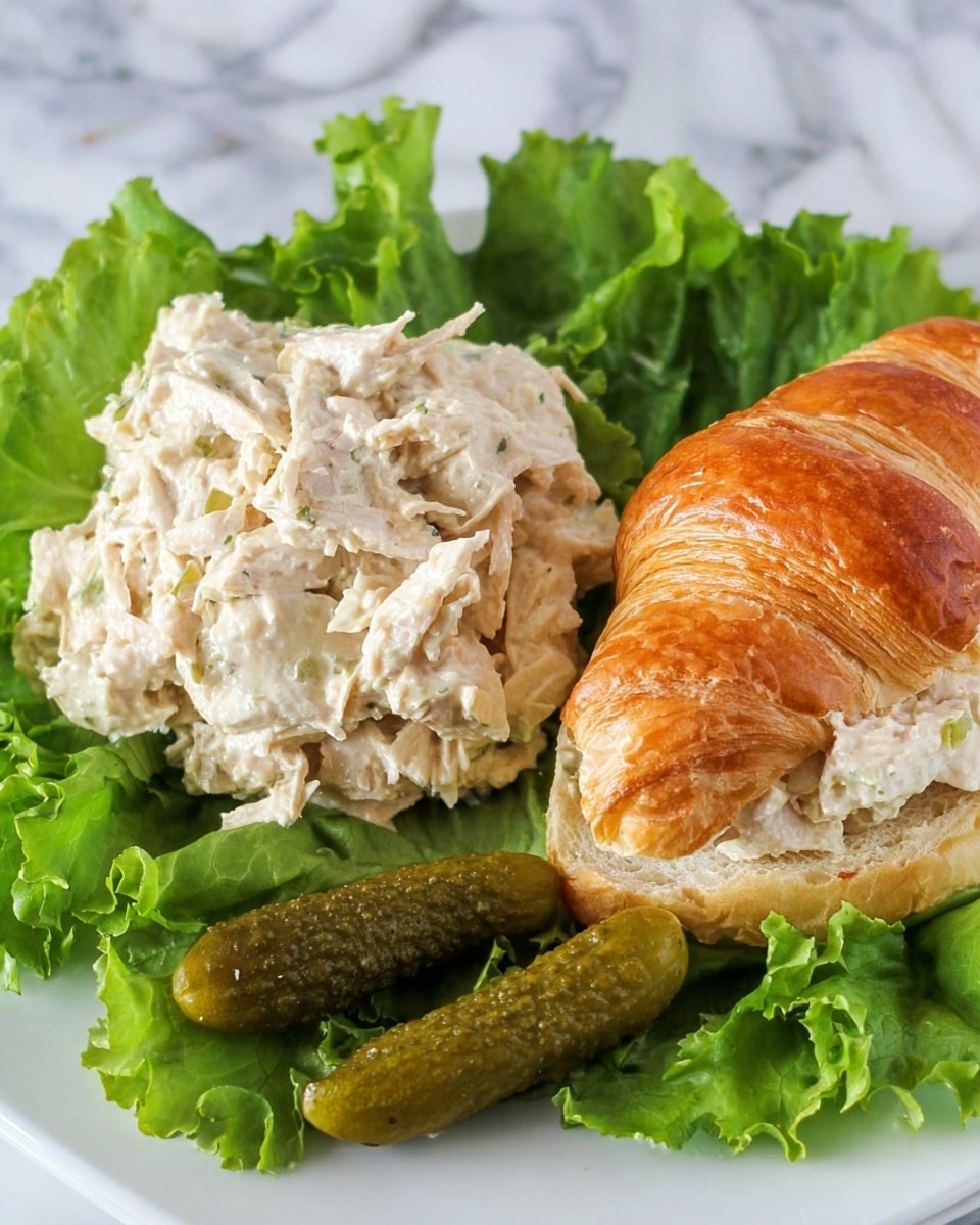 The image shows a white plate with a serving of light beige chicken salad, placed on a bed of dark and bright green leafy lettuce. To the right of the salad is a croissant sandwich filled with the same chicken salad, showing a flaky, golden brown top layer and light soft interior. Below the sandwich, there are several small green pickles resting on more leafy lettuce. The textures highlight the creamy chicken salad, the crispness of the lettuce, the shiny surface of the pickles, and the flaky croissant. The plate is set on a white marbled surface. photo taken with an iphone --ar 4:5 --v 7