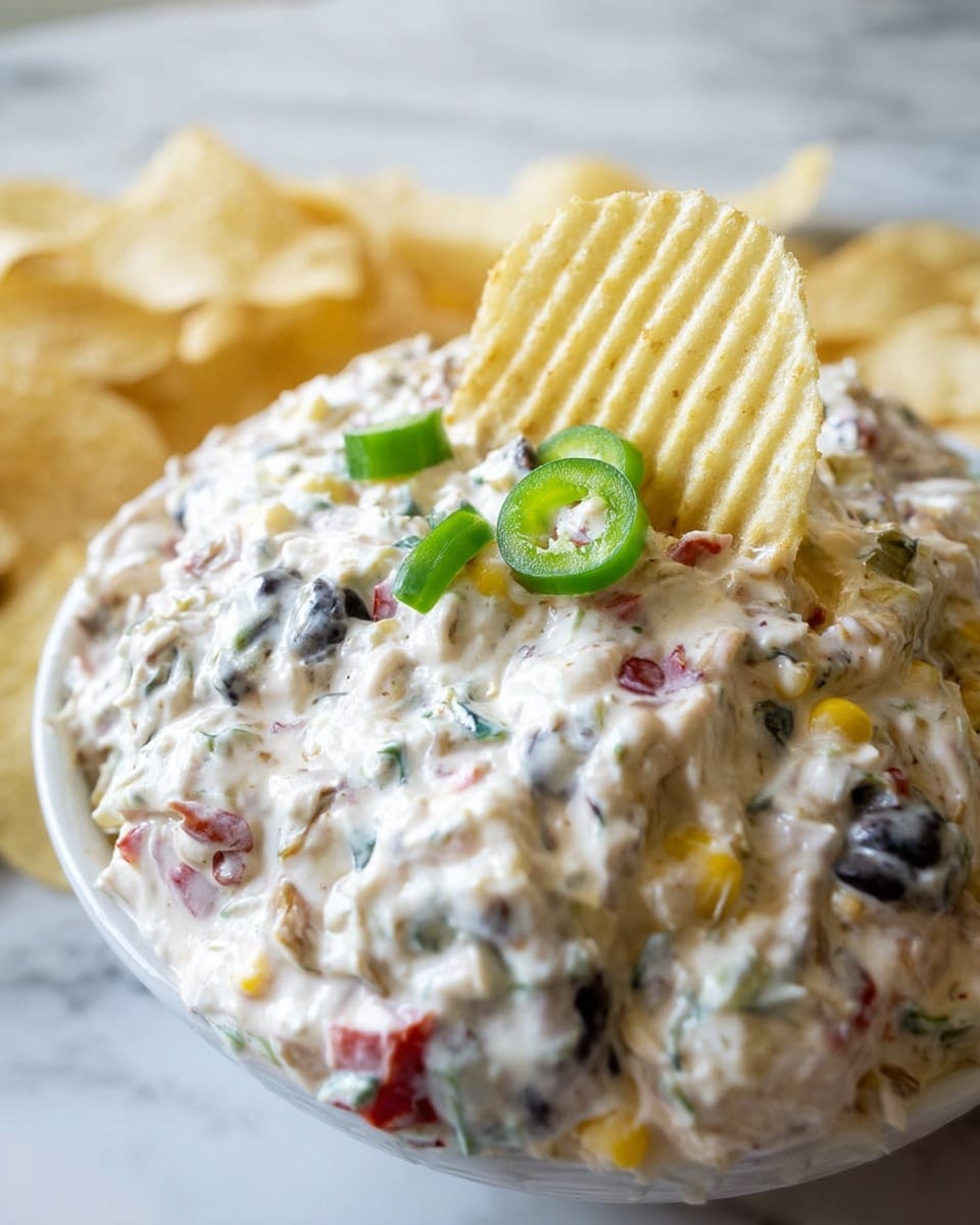 The image shows a close-up of a creamy dip with a thick white layer full of small bits of black olives, red peppers, yellow corn, and green sliced peppers mixed inside. A ridged, pale yellow potato chip is stuck into the dip, sitting near the top center. The dip looks rich and chunky with a smooth texture, and small green pepper slices sit on top, giving a fresh look. In the blurred background, some more chips can be seen resting on a white marbled surface. Photo taken with an iphone --ar 4:5 --v 7