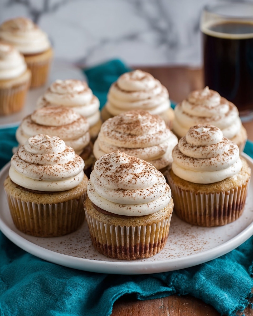 There is a white plate filled with eight cupcakes arranged closely together, each cupcake has two main layers: a light brown cake base at the bottom and a thick swirl of light cream colored frosting on top, shaped in three rounded layers, dusted with brown powder evenly over the frosting. The cupcakes are placed on a teal cloth over a wooden surface, with a white marbled background blurred out. In the back right side, there is a blurry dark beverage in a glass. Photo taken with an iphone --ar 4:5 --v 7