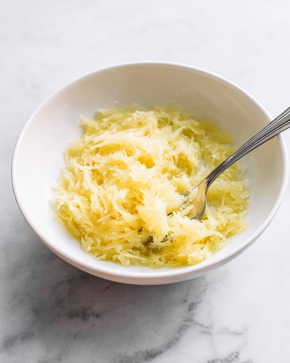 Inside a white bowl, there is one layer of pale yellow grated food with a moist texture that fills the center and spreads toward the edges of the bowl. A silver fork rests halfway inside the grated food, angled with its tines partly covered by the food. The bowl sits on a white marbled surface that adds a soft texture and subtle gray veins in the background. photo taken with an iphone --ar 4:5 --v 7