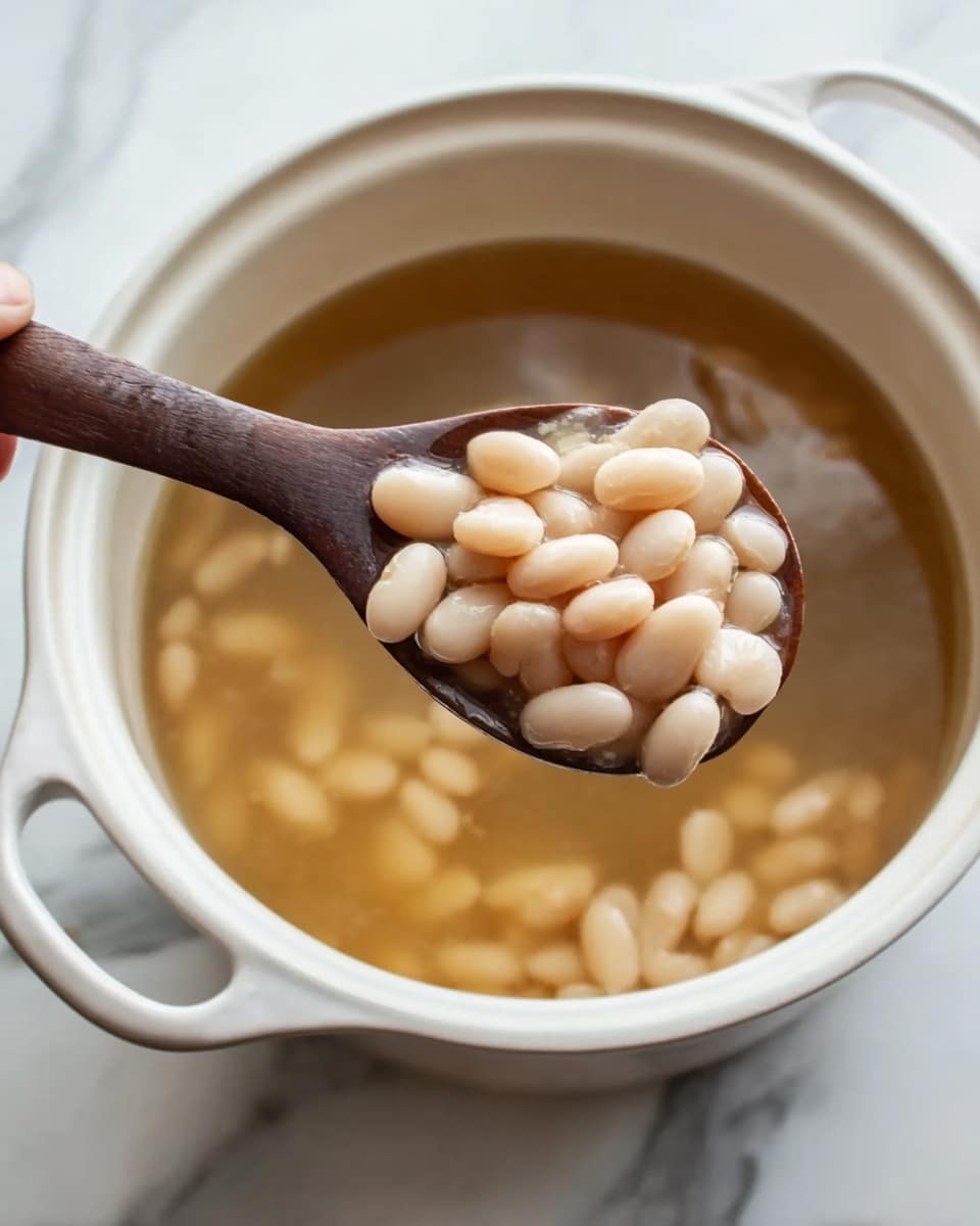 A white pot filled with a light brown broth and many white beans soaking in the liquid. A dark wooden spoon is scooping up a pile of the white, smooth beans from the pot, showing their oval shape and soft texture. The pot is placed on a white marbled surface with subtle gray veins. The image captures the warm, simple look of cooked beans in broth. photo taken with an iphone --ar 4:5 --v 7