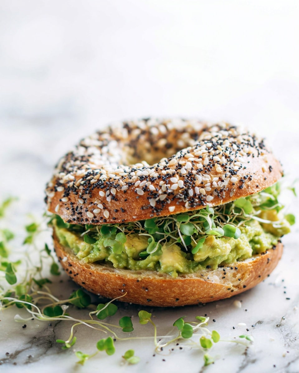 The image shows a sandwich made with a sesame and poppy seed bagel that has a lightly toasted golden-brown color. The top bagel half is sprinkled generously with white sesame seeds and black poppy seeds, adding texture. Inside the sandwich, there is a thick layer of bright green mashed avocado, which looks creamy and fresh. On top of the avocado, there are small fresh green leaves and sprouts peeking out from the edges, adding a light, leafy texture and color contrast. The sandwich sits on a white marbled surface with some scattered green sprouts around it, giving a fresh look. The photo taken with an iphone --ar 4:5 --v 7