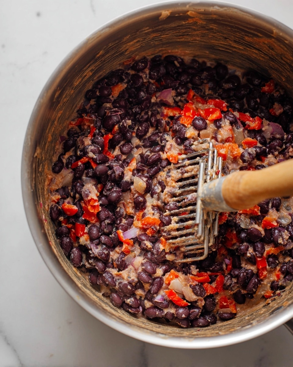 A close-up view inside a pot shows cooked black beans mixed with finely chopped red bell peppers and small pieces of onion. The beans are partially mashed, creating a thick, creamy texture with visible whole and broken beans. The colors are mainly dark purple-black from the beans, bright red from the bell peppers, and pale beige from the mashed beans and onions. A metal potato masher with a wooden handle is partially submerged, pressing down the mixture. The pot's interior is silver with some residue, and the pot sits on a white marbled surface. photo taken with an iphone --ar 4:5 --v 7