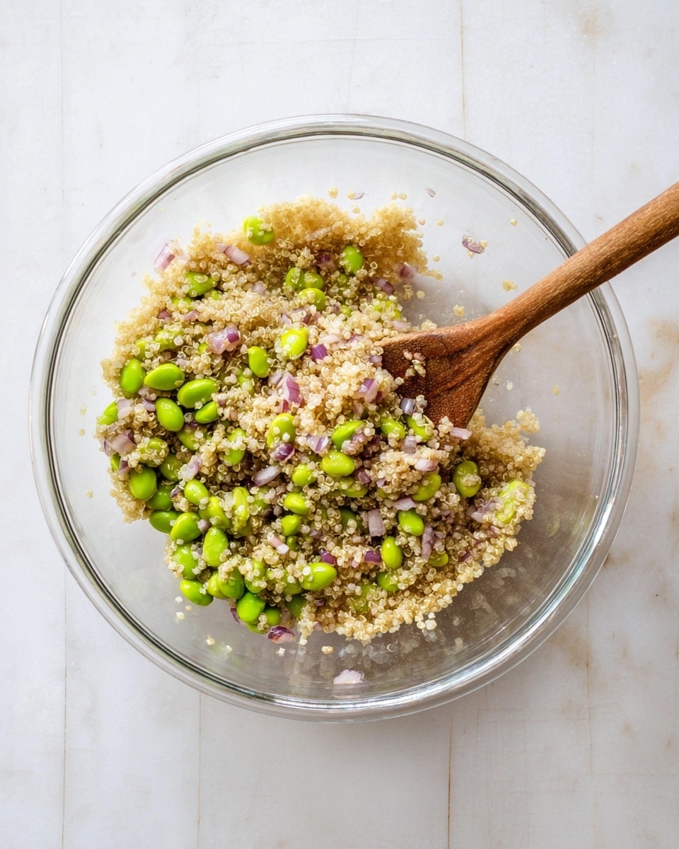 A clear glass bowl holds a mix of light beige quinoa with bright green edamame beans and small pieces of light purple shallots scattered evenly throughout. A wooden spoon rests inside the bowl on the right side, with some quinoa grains stuck to it. The bowl sits on a white marbled surface, giving a clean and fresh look to the scene. photo taken with an iphone --ar 4:5 --v 7