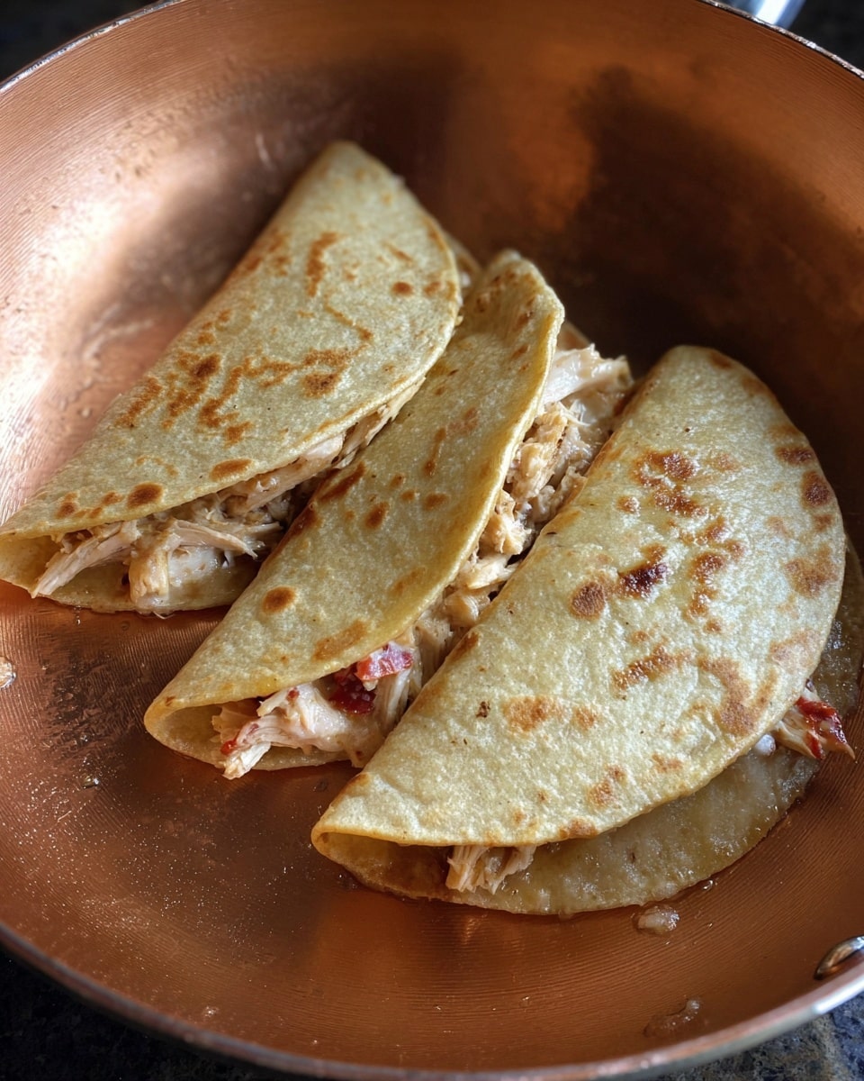 Three small, golden-brown folded tortillas sit inside a copper pan. Each tortilla is filled with light-colored shredded chicken mixed with bits of red pieces, likely tomatoes or peppers, peeking out slightly from the edges. The tortillas have a lightly crisped and bumpy texture on the outside, with some areas showing slight oil glistening. The pan gives a warm background, highlighting the soft yet firm look of the tortillas. There is no additional garnish or sauce visible around the tortillas. photo taken with an iphone --ar 4:5 --v 7