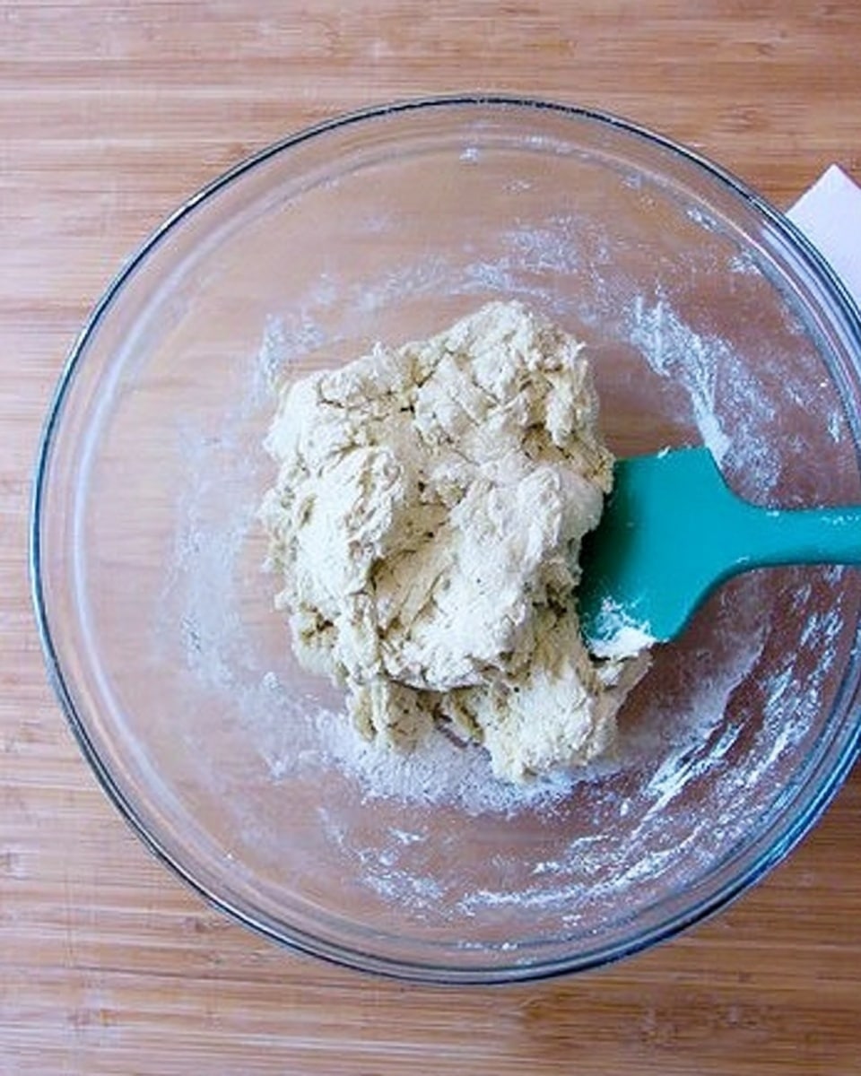 A clear glass bowl holds a rough, uneven ball of pale dough mixed with some flour still visible in and around it, showing a slightly sticky texture. A turquoise spatula rests inside the bowl, with its blade partially covered by the dough on the right side. The bowl sits on a light wooden surface, adding contrast to the soft cream and white tones of the dough and flour. photo taken with an iphone --ar 4:5 --v 7