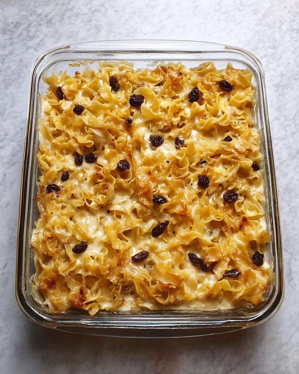 The image shows a glass baking dish with a baked noodle casserole. The top layer is golden yellow noodles, slightly browned at the edges, with some dark raisins scattered across. The noodles look soft and are loosely arranged, with creamy white sauce visible between them in places. The dish is placed on a white marbled surface. Photo taken with an iphone --ar 4:5 --v 7
