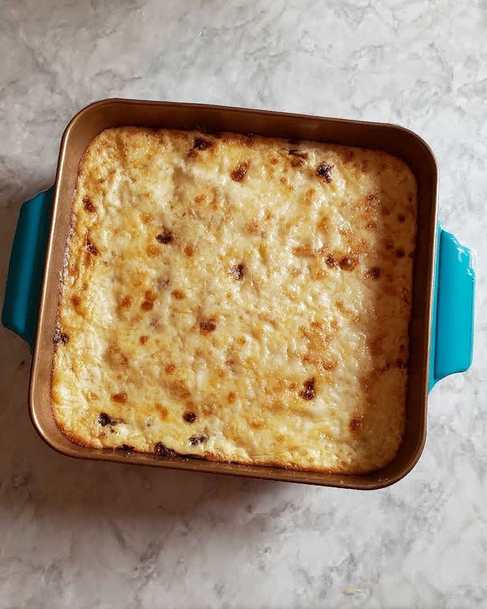 A square pan filled with a baked dish that has a light golden brown top layer, dotted with small browned spots and some scattered darker bits. The texture of the top looks soft and slightly uneven, indicating a creamy or cheesy mixture beneath. The pan is placed on a white marbled surface with a small turquoise handle cover on the right side. photo taken with an iphone --ar 4:5 --v 7