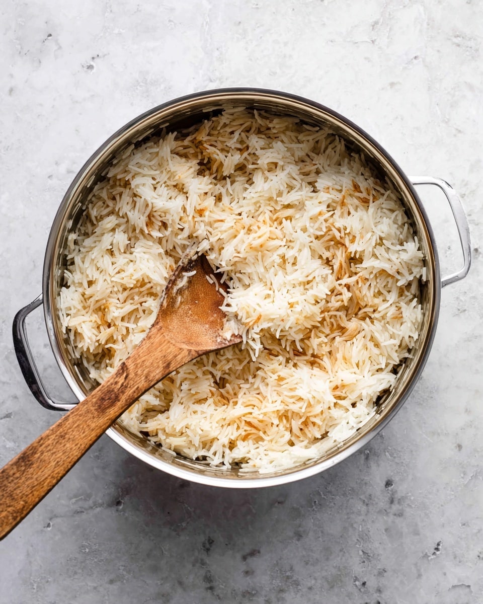 A silver pot filled with cooked white rice that has some light brown toasted parts mixed in; the rice grains are long and fluffy with a slightly dry texture. A wooden spoon with a smooth, flat surface rests inside the pot, positioned diagonally from the bottom left to the top right. The pot is placed on a white marbled surface with faint grey and black speckles. photo taken with an iphone --ar 4:5 --v 7