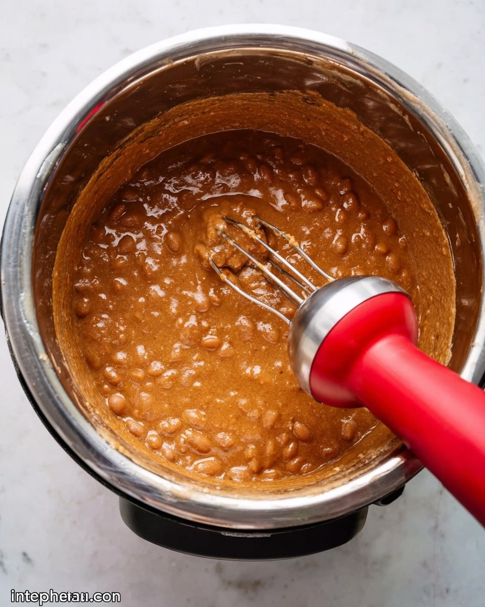 The image shows a top view of a silver metal pot with thick brown bean stew inside. The bean stew looks creamy and slightly chunky with a shiny wet surface. A red and silver handheld mixer is partially inside the pot, blending the beans. The pot is sitting on a white marbled surface. photo taken with an iphone --ar 4:5 --v 7