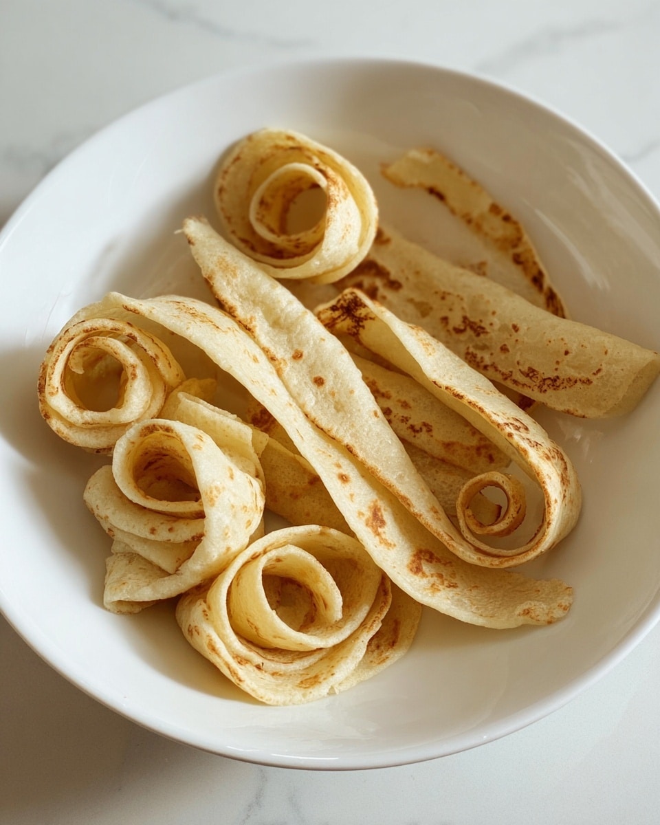A white bowl holds several light beige spirals that resemble thinly rolled pieces of flatbread or pancake. Each spiral is shaped loosely into curls, showing some darker browned edges and spots on the outer side, giving a toasted look. The texture appears soft but with slight firmness on the browned areas. The bowl sits on a white marbled surface, with no other items around. Photo taken with an iphone --ar 4:5 --v 7