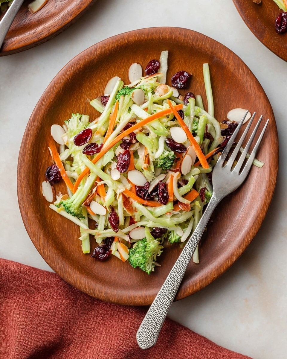 A wooden plate holds a bright salad made of thin green strips of broccoli stems, orange carrot sticks, white almond slices scattered on top, and dark red dried cranberries mixed throughout, giving a fresh and crunchy look. The salad has a light coating of dressing that adds a soft shine. A silver fork with a textured handle rests on the right side of the plate. The plate sits on a white marbled surface with a hint of a rust-colored cloth napkin nearby. Photo taken with an iphone --ar 4:5 --v 7