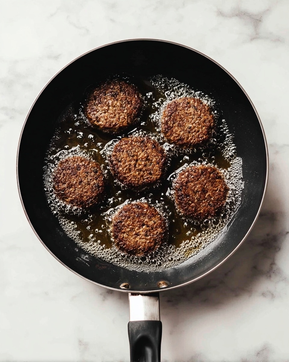 A black frying pan with a black handle holds six dark brown patties cooking in bubbling oil. The patties have a coarse texture with small light-colored spots and were arranged in a roughly circular shape inside the pan. The pan sits on a white marbled surface with light gray veins visible. photo taken with an iphone --ar 4:5 --v 7