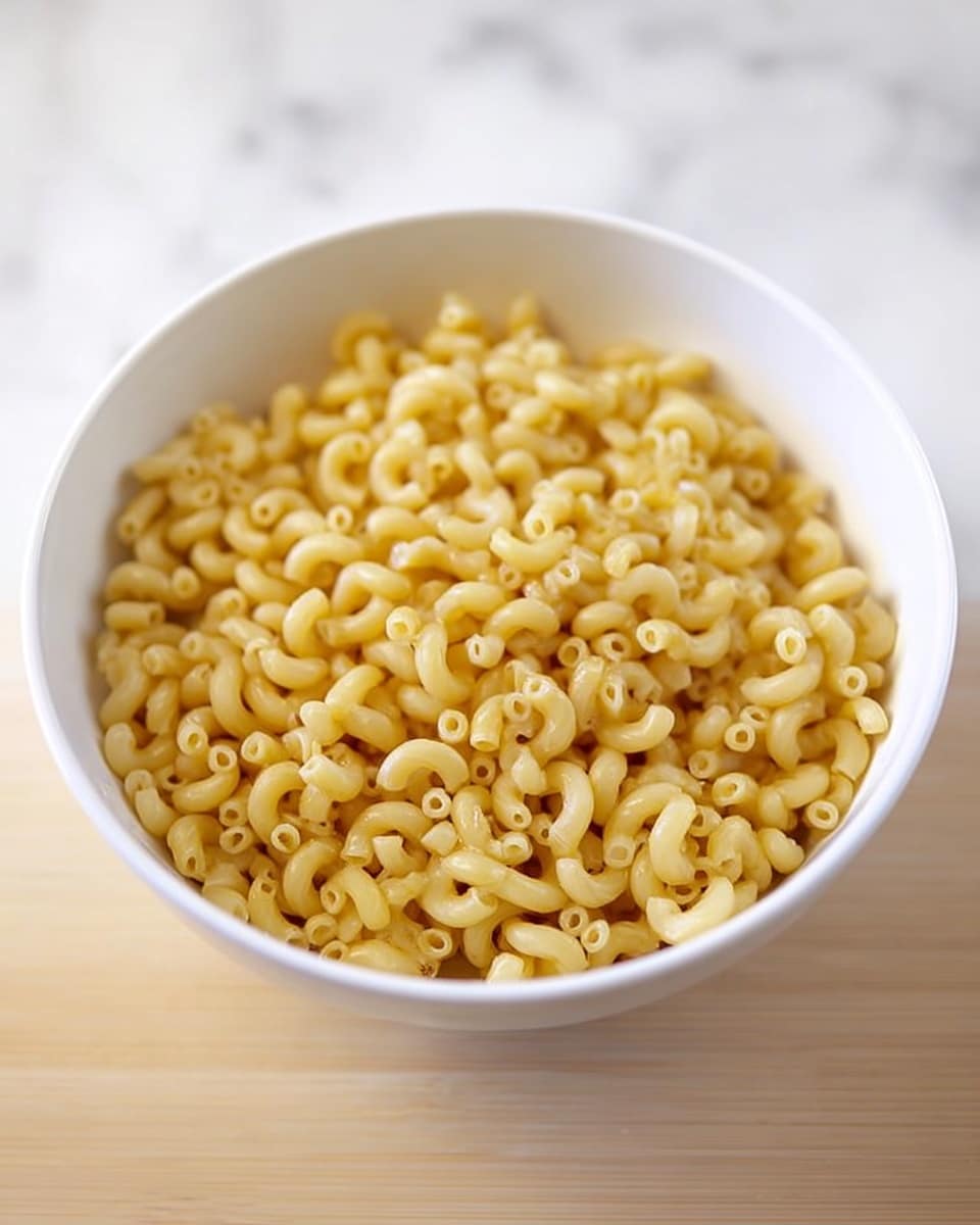 A white bowl filled with small, uncooked elbow macaroni pasta, sitting on a light wooden surface. The pasta has a smooth texture and a light yellow color, tightly packed in the bowl that is shown from a slightly above angle. The background is a white marbled texture. photo taken with an iphone --ar 4:5 --v 7