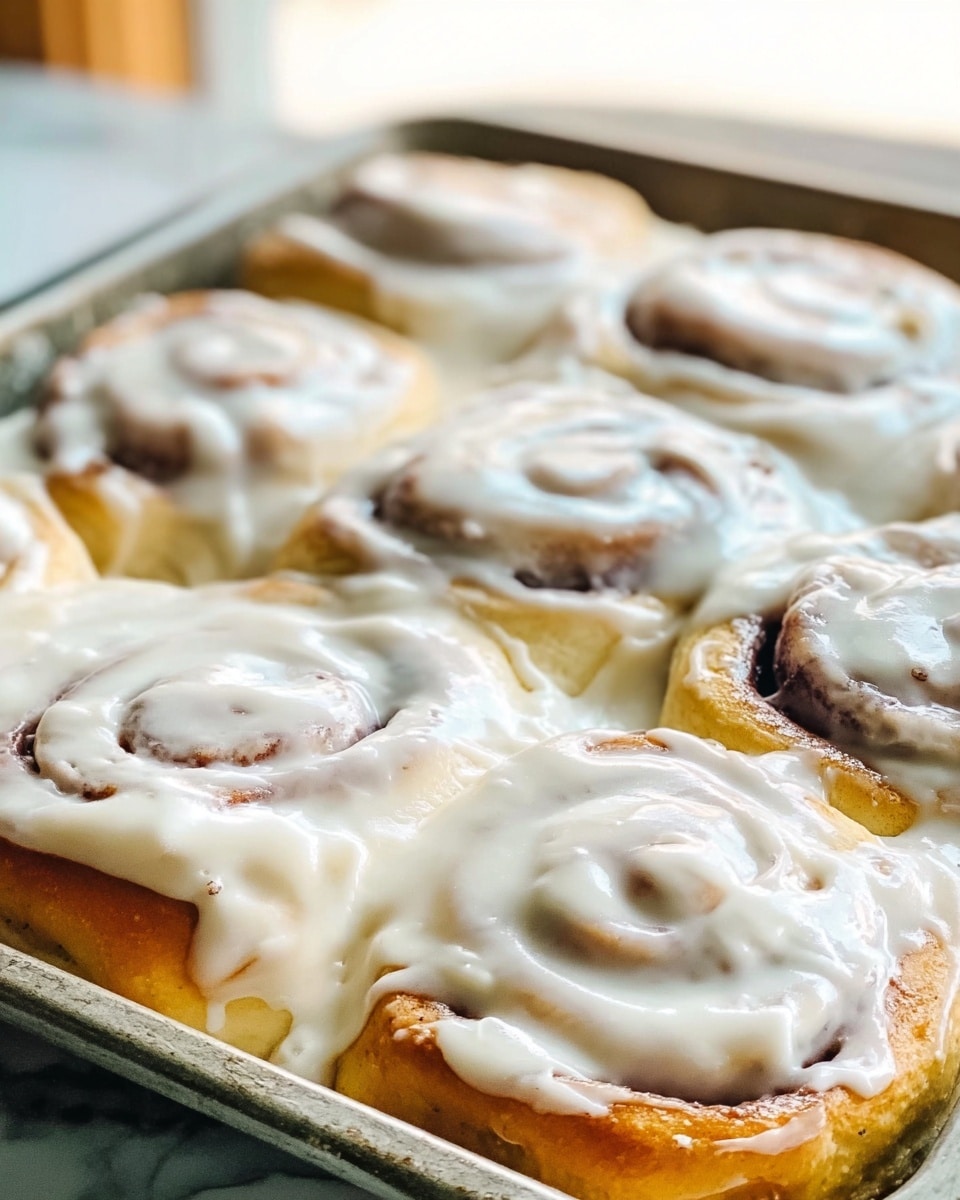 A close-up view of a baking tray filled with a single layer of golden brown cinnamon rolls covered thickly with creamy white icing that looks smooth and spread unevenly, with the rolls arranged closely together showing swirls of light brown cinnamon inside. The tray rests on a white marbled surface with soft, natural lighting making the icing look shiny and fresh. photo taken with an iphone --ar 4:5 --v 7