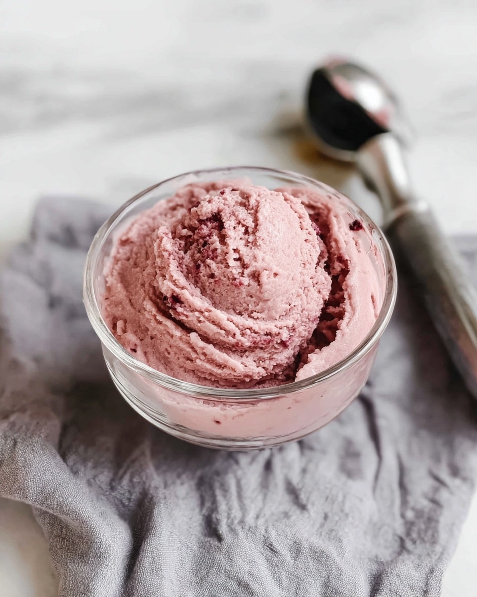 A clear glass bowl holds a single layer of thick, creamy ice cream in a dusty pink color with a slightly textured surface showing swirls and small bits mixed in. The bowl is placed on a soft, crumpled gray cloth that rests on a white marbled surface. Nearby, a silver ice cream scoop with a black handle lies slightly out of focus, suggesting recent use. The scene is lit softly, highlighting the smooth and slightly rough texture of the ice cream. photo taken with an iphone --ar 4:5 --v 7