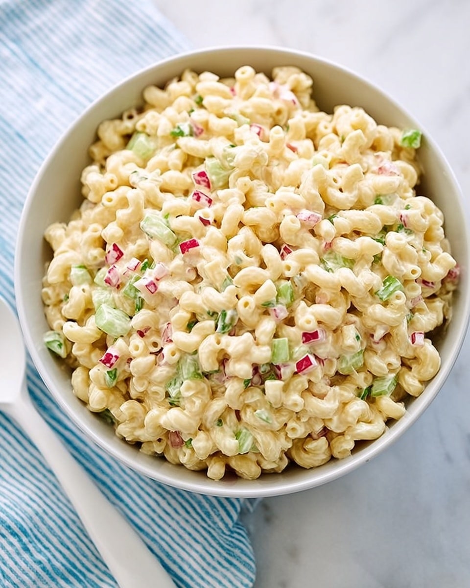 The image shows a white bowl full of creamy macaroni salad made with small elbow pasta. The salad has a shiny creamy texture and is mixed with small pieces of green celery and red radish scattered evenly throughout. The bowl sits on a white marbled surface with a white spoon and a striped white and blue cloth partially visible nearby. The colors are soft with the pale yellow pasta contrasting gently with the green and red bits in the mix, giving a fresh and light look. photo taken with an iphone --ar 4:5 --v 7
