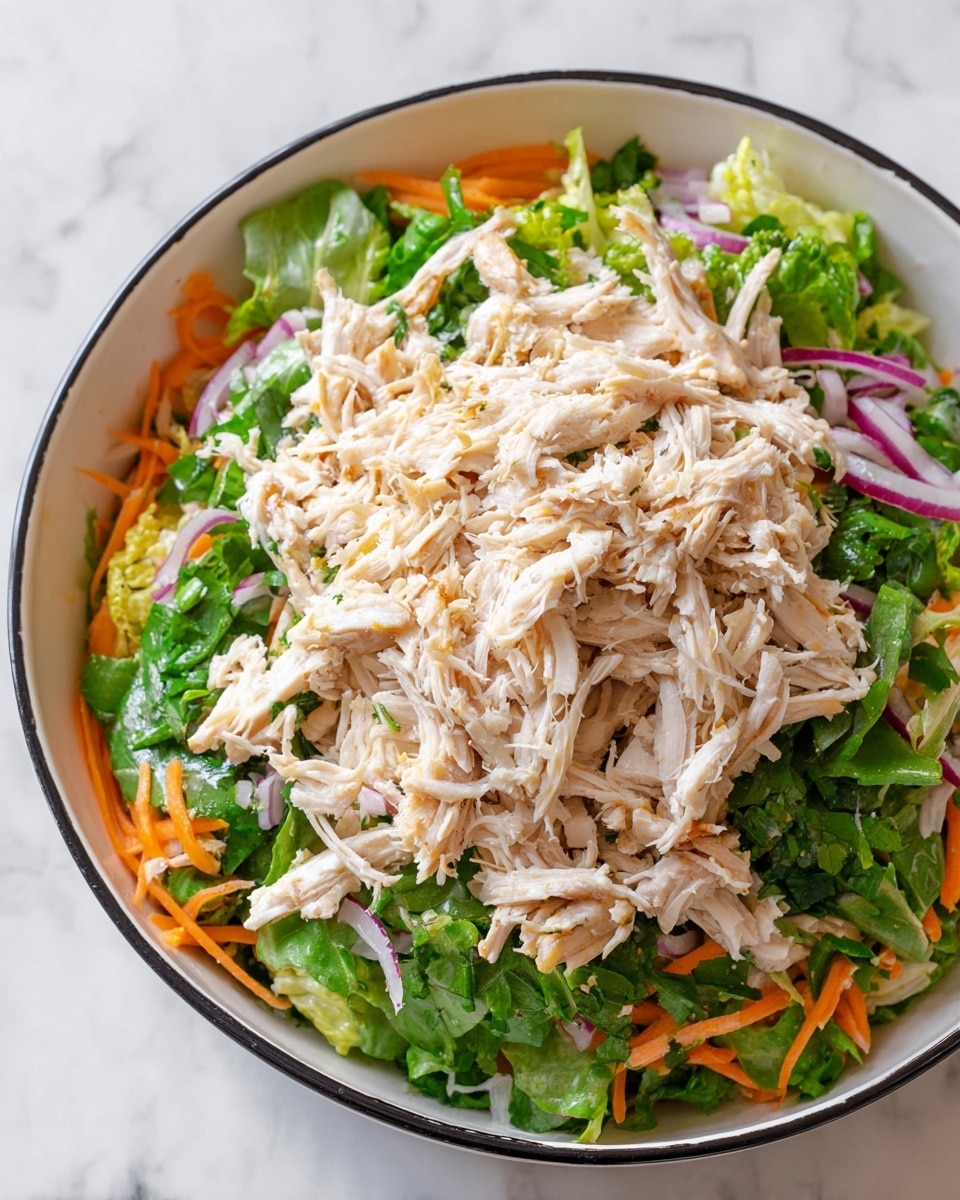 A white bowl with a black rim holds a layered salad on a white marbled surface. The bottom layer is a mix of bright green leafy herbs and lettuce, with thin strips of vibrant orange carrots scattered around. Thinly sliced purple onions are mixed in with the greens. On top, there is a large mound of light beige shredded chicken, spread out evenly to cover the vegetables below. The colors contrast strongly, showing freshness and texture in each layer, with the shredded chicken’s soft strands placed loosely over the crisp colorful vegetables photo taken with an iphone --ar 4:5 --v 7