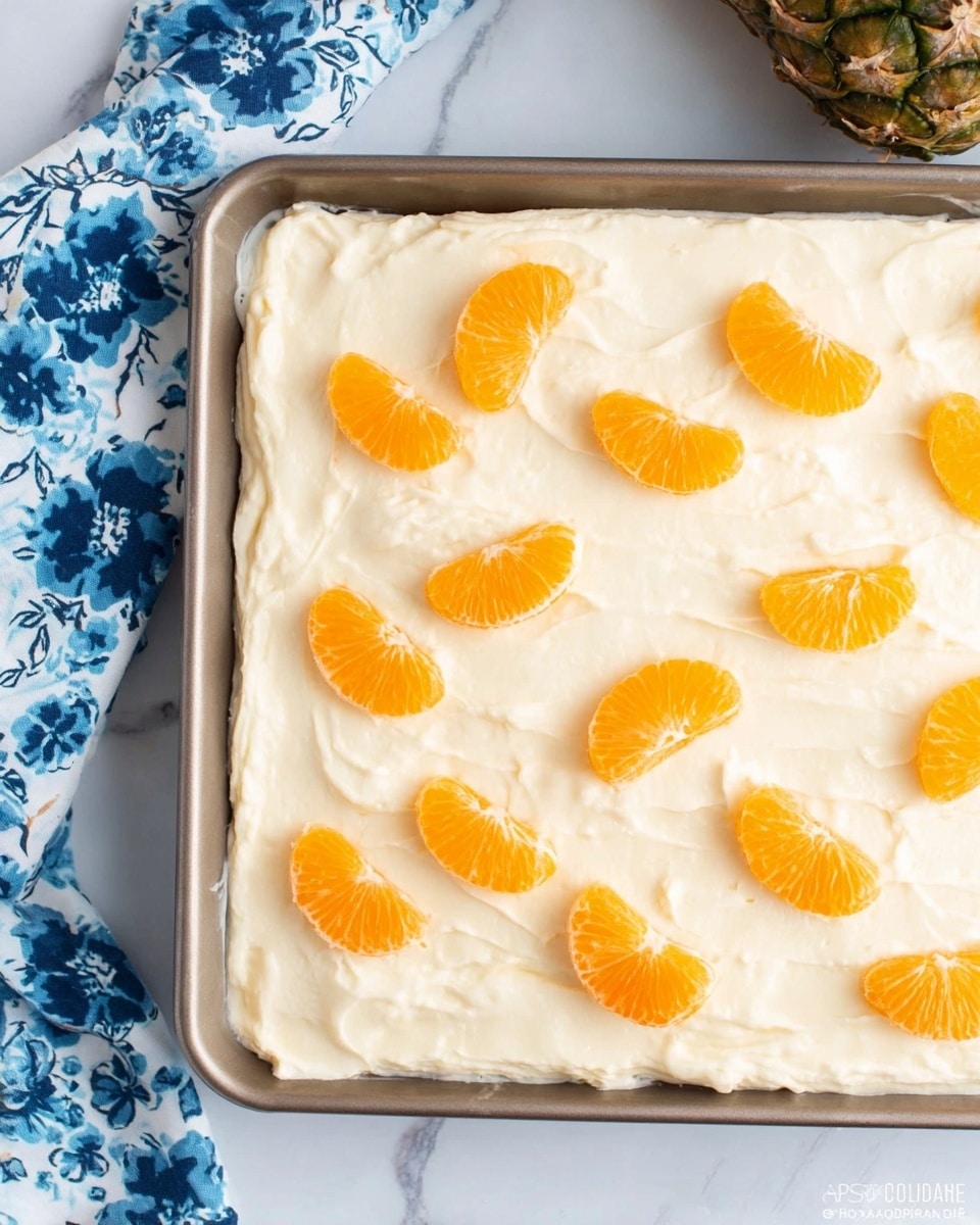 There is a large rectangular tray filled with a thick, creamy white layer spread evenly across the surface. On top, there are pairs of bright orange mandarin segments arranged in two rows, each pair sitting close together and spaced evenly apart. The tray rests on a white marbled surface with a piece of cloth featuring blue flowers on the upper left side and a small part of a pineapple crown visible in the upper right corner. The texture of the creamy layer is smooth but slightly uneven, showing soft ridges and swirls where it has been spread. photo taken with an iphone --ar 4:5 --v 7