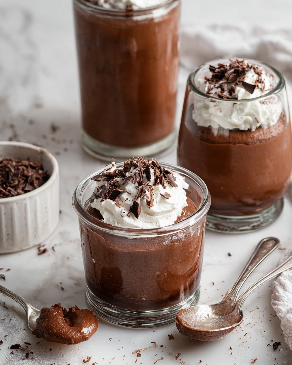 Four small glass jars each hold two visible layers: a smooth dark brown chocolate pudding base topped with a thick white whipped cream layer. The whipped cream is decorated with dark chocolate shavings scattered unevenly on top. The jars sit on a white marbled surface with some small chocolate shavings scattered around, alongside two vintage silver spoons and a small white bowl filled with more chocolate shavings. One jar rests on a light brown ceramic coaster. The scene is evenly lit with a clean and minimal look. photo taken with an iphone --ar 4:5 --v 7