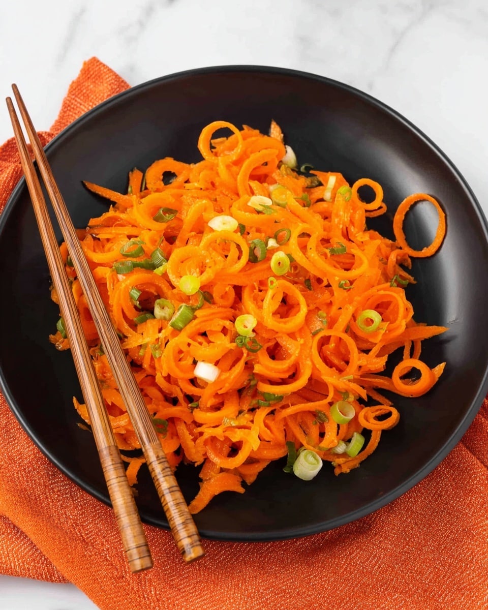 The image shows a white plate with a black bowl on top filled with bright orange curly strips of carrot mixed with small pieces of green onion scattered throughout. The carrot curls create a layered, spiral texture that covers most of the bowl's surface. A pair of wooden chopsticks rests diagonally across the bowl. The bowl and chopsticks sit on a white marbled surface with an orange cloth in the background. Photo taken with an iphone --ar 4:5 --v 7