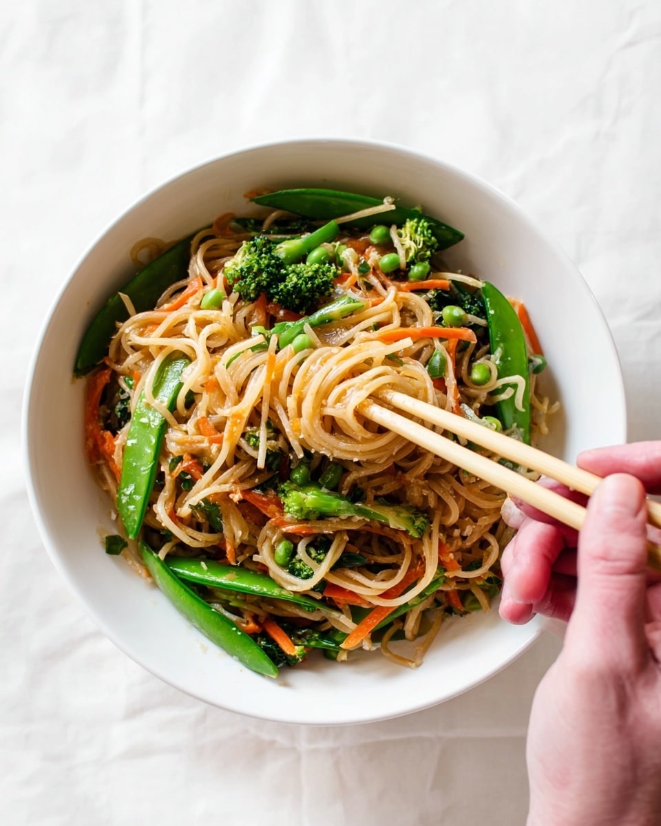 A white bowl filled with a colorful noodle dish is shown on a white marbled surface. The bottom layer consists of light brown, thin noodles with a slightly glossy texture. Mixed in are bright green snap peas and small broccolini pieces, adding a crisp, fresh look. Orange carrot strips are scattered throughout, creating a contrast of color. A woman's hand holding pale yellow chopsticks reaches into the bowl, picking up noodles with vegetables. The overall image has a fresh and vibrant appearance, highlighting the mix of textures and colors. Photo taken with an iphone --ar 4:5 --v 7