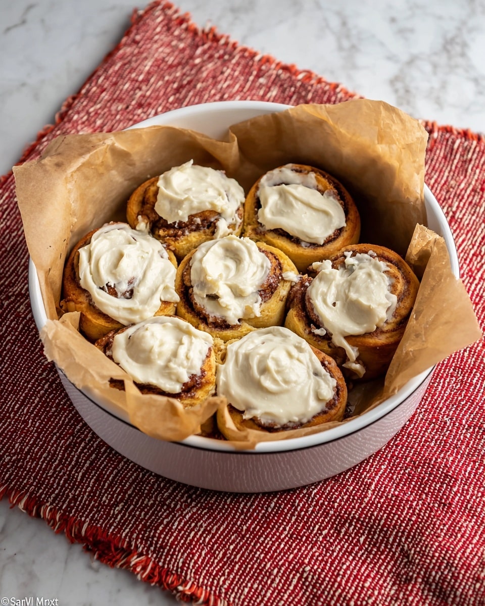 A single cinnamon roll sits on a white round plate, showing one main layer of golden brown dough spiraled with darker cinnamon filling and topped with a swirl of creamy white icing slightly dripping down the sides. The roll has a soft, textured surface with some shiny spots from the icing. Next to the roll is a silver fork resting on the plate. In the background on a white marbled surface, there is a round white baking dish lined with light brown parchment paper holding several similar cinnamon rolls covered with white icing. The scene has a warm and cozy feel with a red patterned cloth under the plate and dish. Photo taken with an iphone --ar 4:5 --v 7