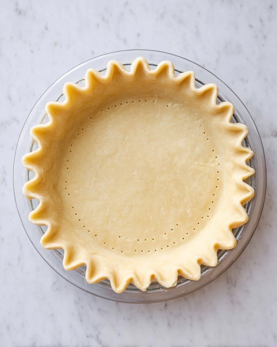 A clear glass pie dish holds a single layer of pale yellow dough pressed evenly to cover the base and sides. The dough has a gently crimped edge all around, creating a wavy border. Small, evenly spaced fork holes are dotted across the flat bottom surface of the dough. This setup sits on a white marbled surface, giving a clean and simple look. photo taken with an iphone --ar 4:5 --v 7
