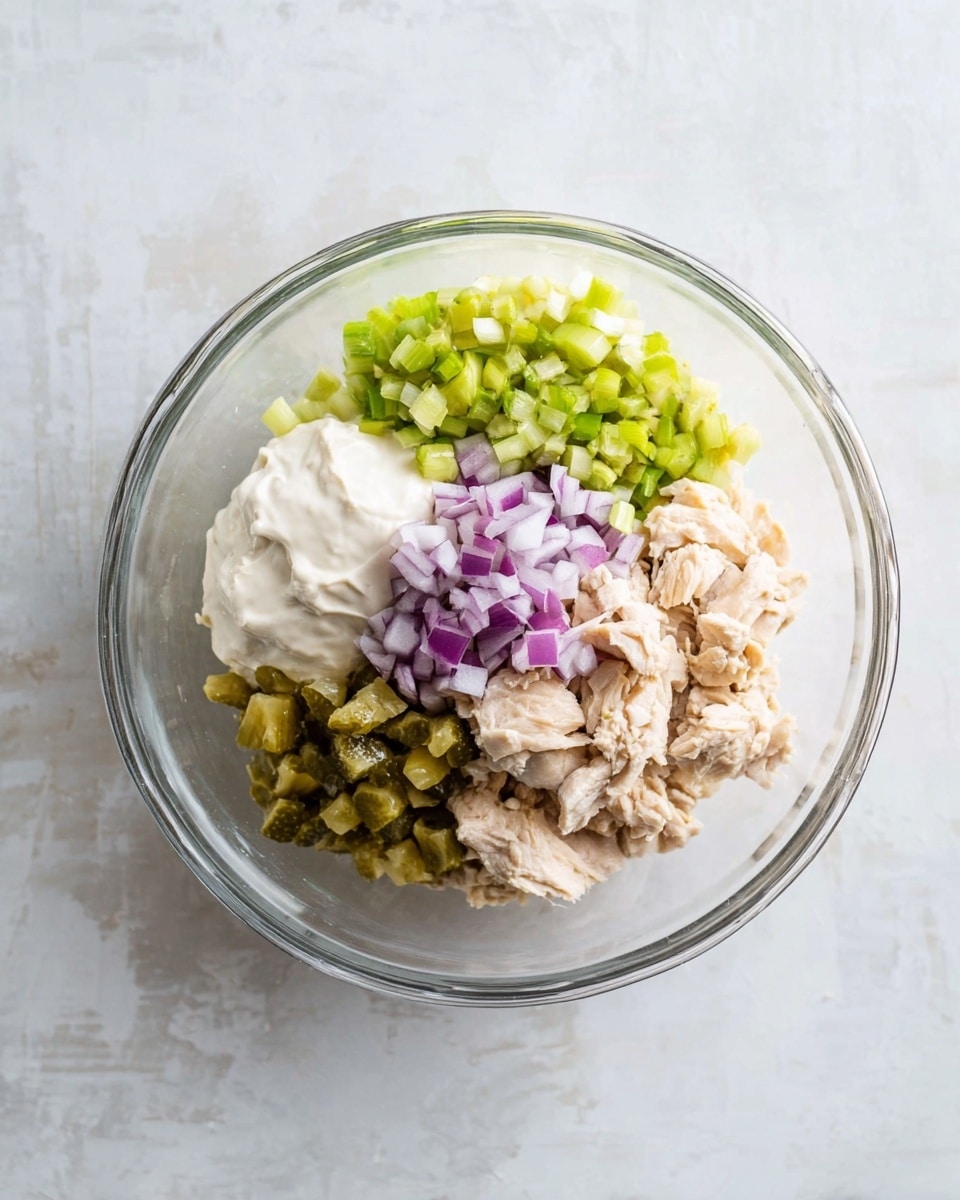 A clear glass bowl sits on a white marbled surface, holding five separate layers of ingredients. There are small pieces of light beige cooked chicken on the bottom right side, a light green chopped celery pile on the top left, finely chopped purple onions on the bottom left, a dollop of creamy white mayonnaise on the top right, and small chopped green pickles just below the celery and onions. Each ingredient is neatly placed and not yet mixed. Photo taken with an iphone --ar 4:5 --v 7