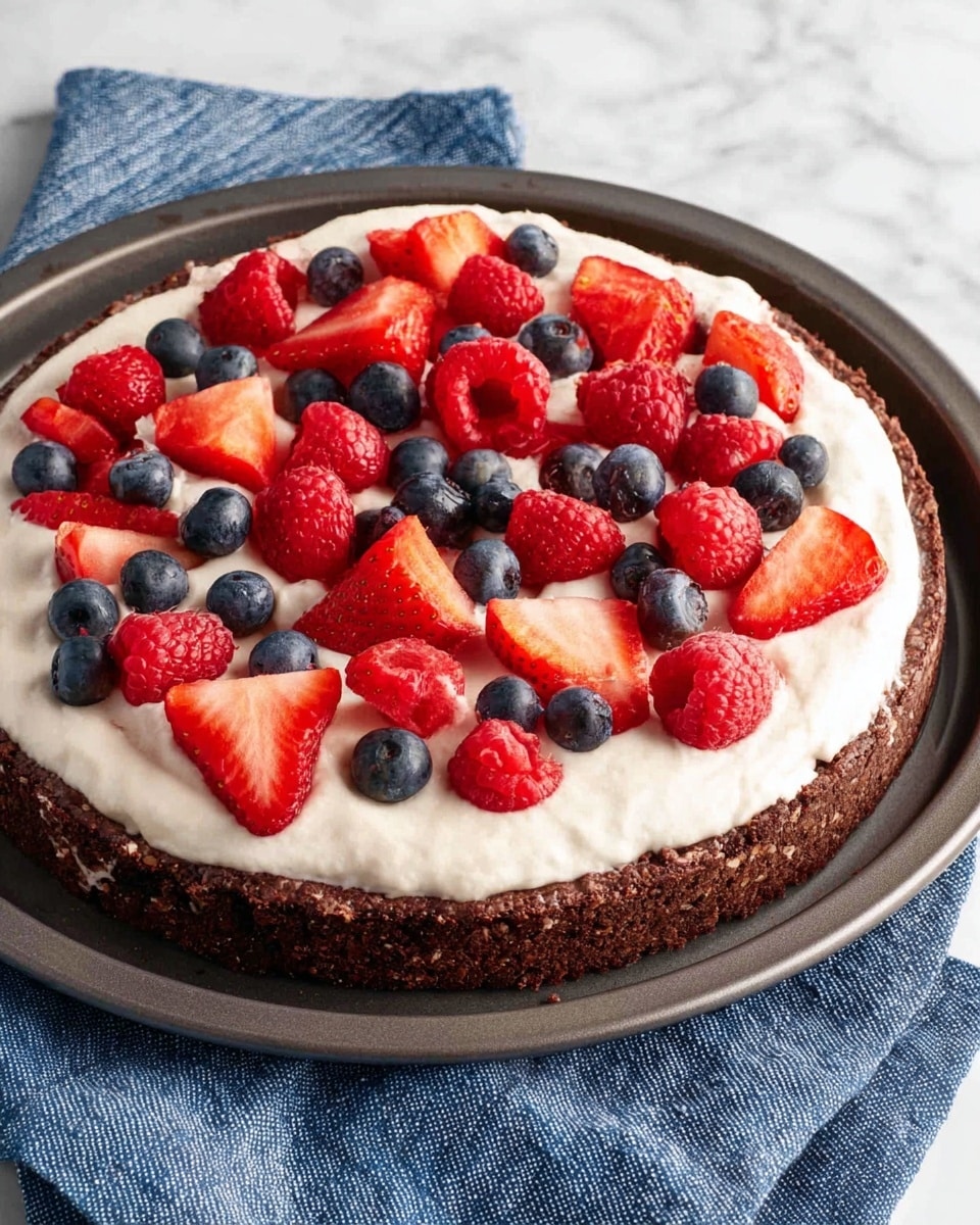 A round dessert with three layers is shown on a dark baking pan resting on a blue cloth over a white marbled surface. The bottom layer is a deep brown, textured chocolate crust. On top of that is a thick, even spread of smooth white cream. Scattered on the cream are fresh red strawberry slices, whole red raspberries, and small dark blue blueberries, adding bright pops of color and texture. The whole scene is well-lit, showing clear details of the dessert. photo taken with an iphone --ar 4:5 --v 7