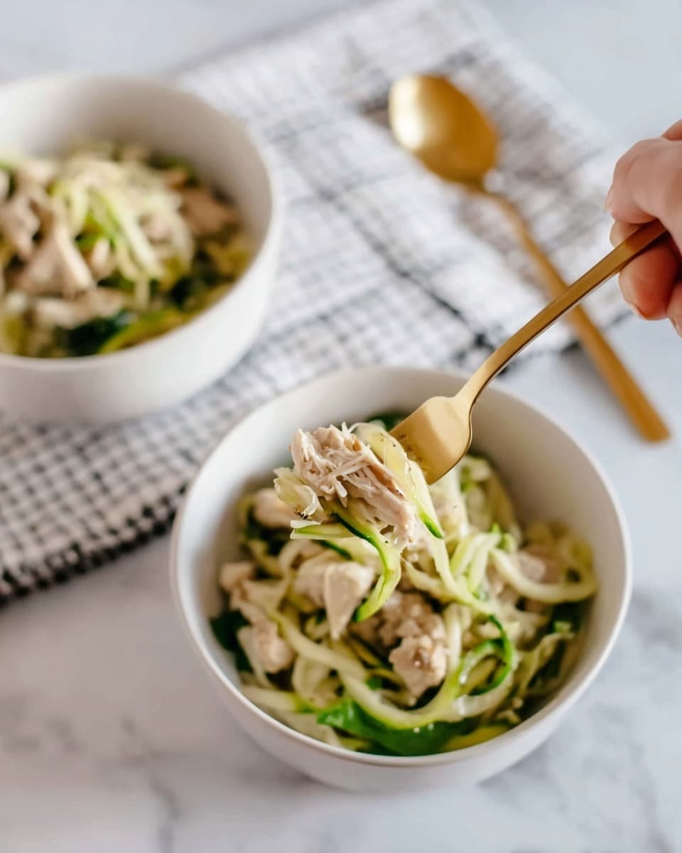 The image shows two white bowls on a white marbled surface, each filled with layers of food. The bottom layer consists of thinly sliced light green zucchini noodles mixed with some leafy green vegetables. On top of the zucchini noodles, there are small pieces of cooked light beige chicken meat. In the foreground, a gold fork held by a woman's hand lifts some of the zucchini noodles and a piece of chicken from one bowl. Behind the bowls, a gold spoon rests on a white cloth with a simple black grid pattern. The lighting is soft, giving the scene a fresh, clean look. photo taken with an iphone --ar 4:5 --v 7