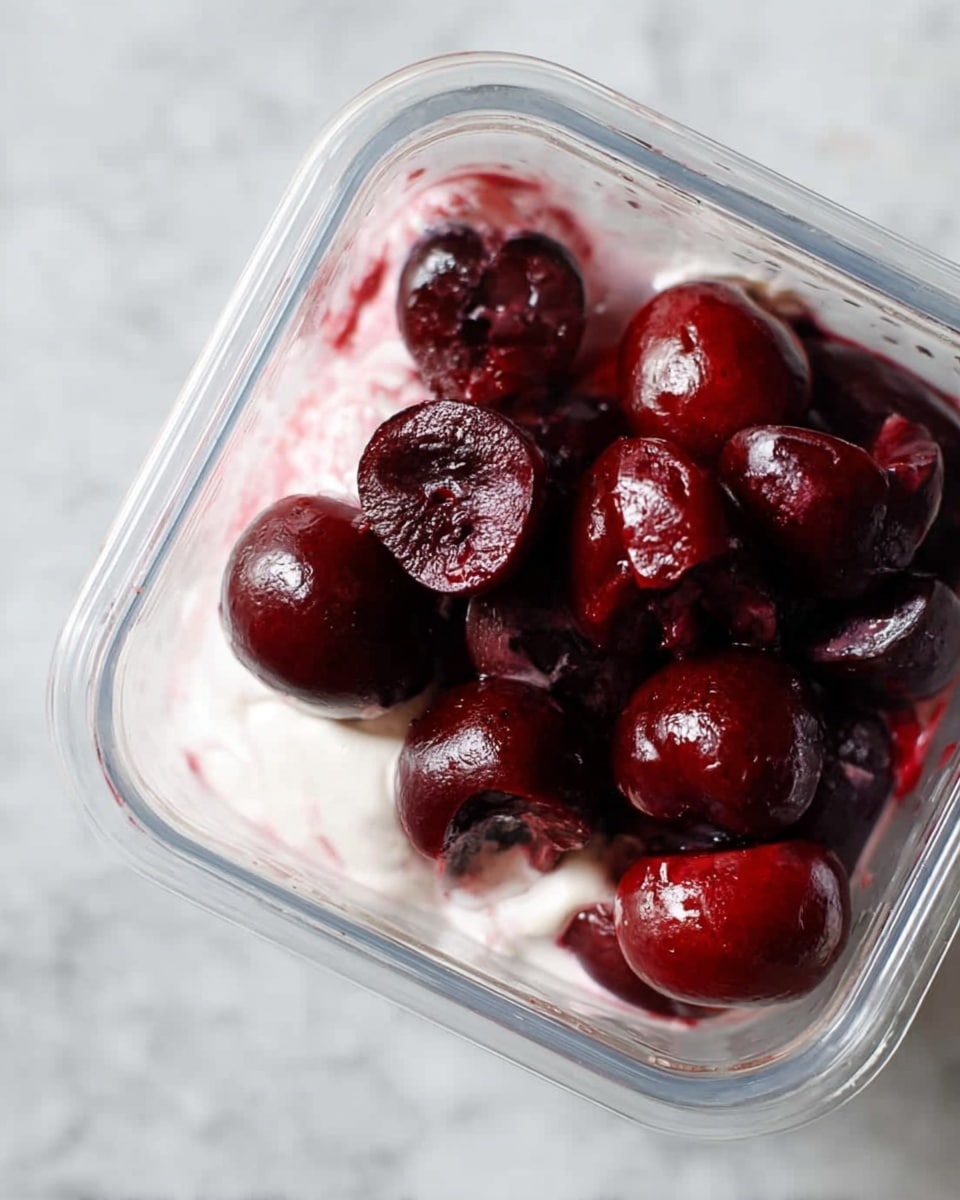 The image shows a close-up view inside a clear square container with a layer of white creamy substance at the bottom and dark red cherry halves piled on top. The cherries have a shiny, moist surface, and some have split open showing their inside. The creamy layer has a soft, smooth texture with a hint of pink from the cherry juice blending in. The scene is set on a white marbled texture background. Photo taken with an iphone --ar 4:5 --v 7