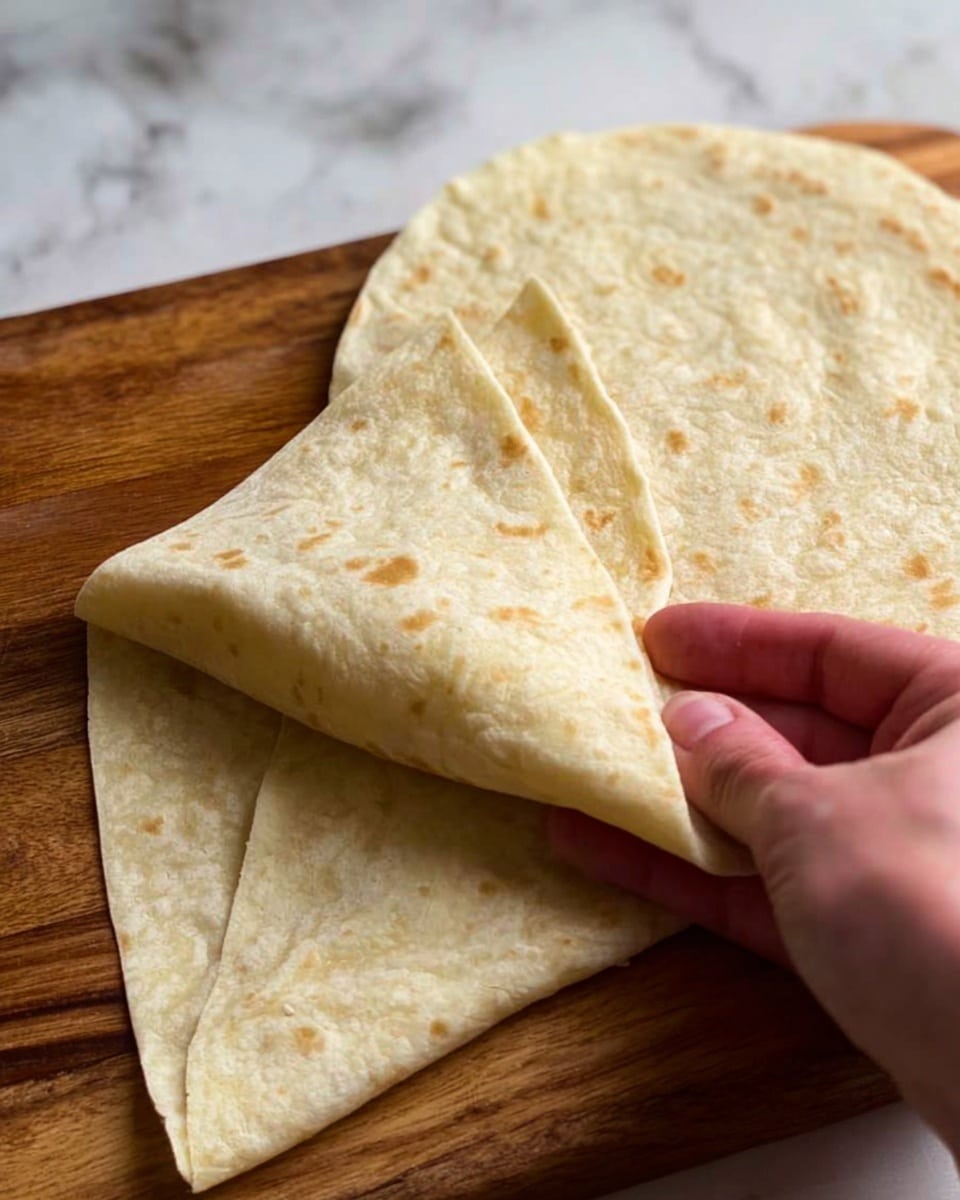 The image shows two large, soft, pale beige tortillas placed on a wooden surface. One tortilla is placed flat on the surface, while a woman's hand is gently lifting or folding the edge of the other tortilla to reveal its thin, flexible texture. The tortillas appear smooth with a slightly uneven surface, showing faint toasted marks and a soft, light texture. The background is a white marbled texture. photo taken with an iphone --ar 4:5 --v 7