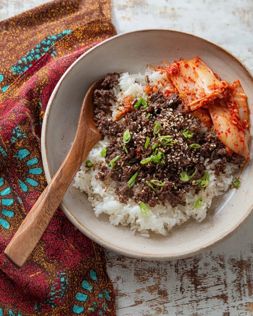 A white round bowl sits on a white marbled surface with a wooden spoon inside. The bottom layer is white rice, topped with a layer of dark brown cooked beef sprinkled with white sesame seeds and small green onion slices. On one side, there is a small portion of bright orange-red kimchi with visible chili flakes. The bowl is partly covered by a colorful cloth with brown, red, yellow, and turquoise patterns. Photo taken with an iphone --ar 4:5 --v 7