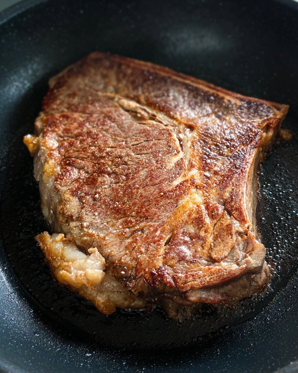 A large single piece of steak is cooking in a black pan, showing a browned surface with a textured, slightly crispy crust. The steak has a light sear with some areas darker brown and others a lighter tan, with visible marbling lines and a few light seasoning specks on top. The pan surface is black and shows some cooking oil around the edges of the steak, creating a slight shine. The background is out of focus but appears to be the surface of the black pan. photo taken with an iphone --ar 4:5 --v 7