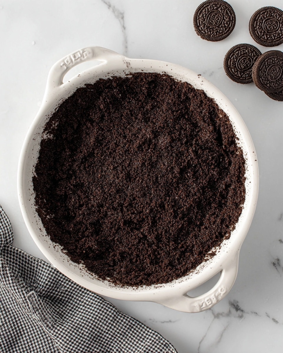The image shows a close-up of a single-layer Oreo crumb crust in a white ceramic pie dish with handles on each side. The crust is dark brown and has a crumbly, rough texture. It evenly fills the bottom and sides of the dish, with slightly uneven edges pressed into the dish shape. The pie dish sits on a white marbled surface, with four whole Oreo cookies placed to the upper right side of the dish, and a folded black and white checkered cloth in the lower left corner. Photo taken with an iphone --ar 4:5 --v 7