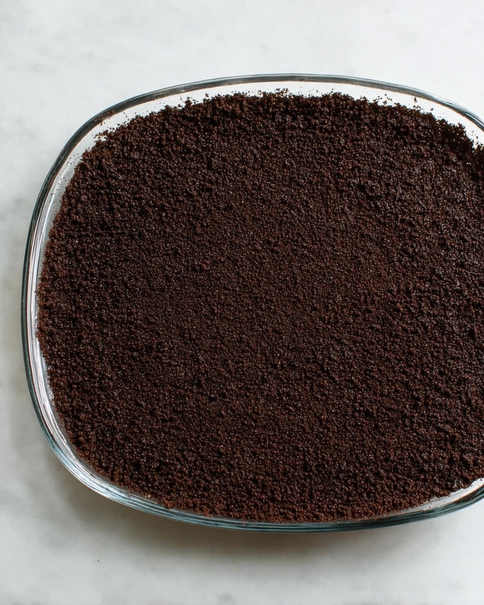 A single layer of dark brown, finely crushed cookie crumbs spread evenly across a clear glass pie dish, creating a smooth, slightly textured base with small granules visible. The dish is placed on a white marbled surface, and the cookie crumb layer covers the entire bottom completely, forming a thin, uniform crust. photo taken with an iphone --ar 4:5 --v 7