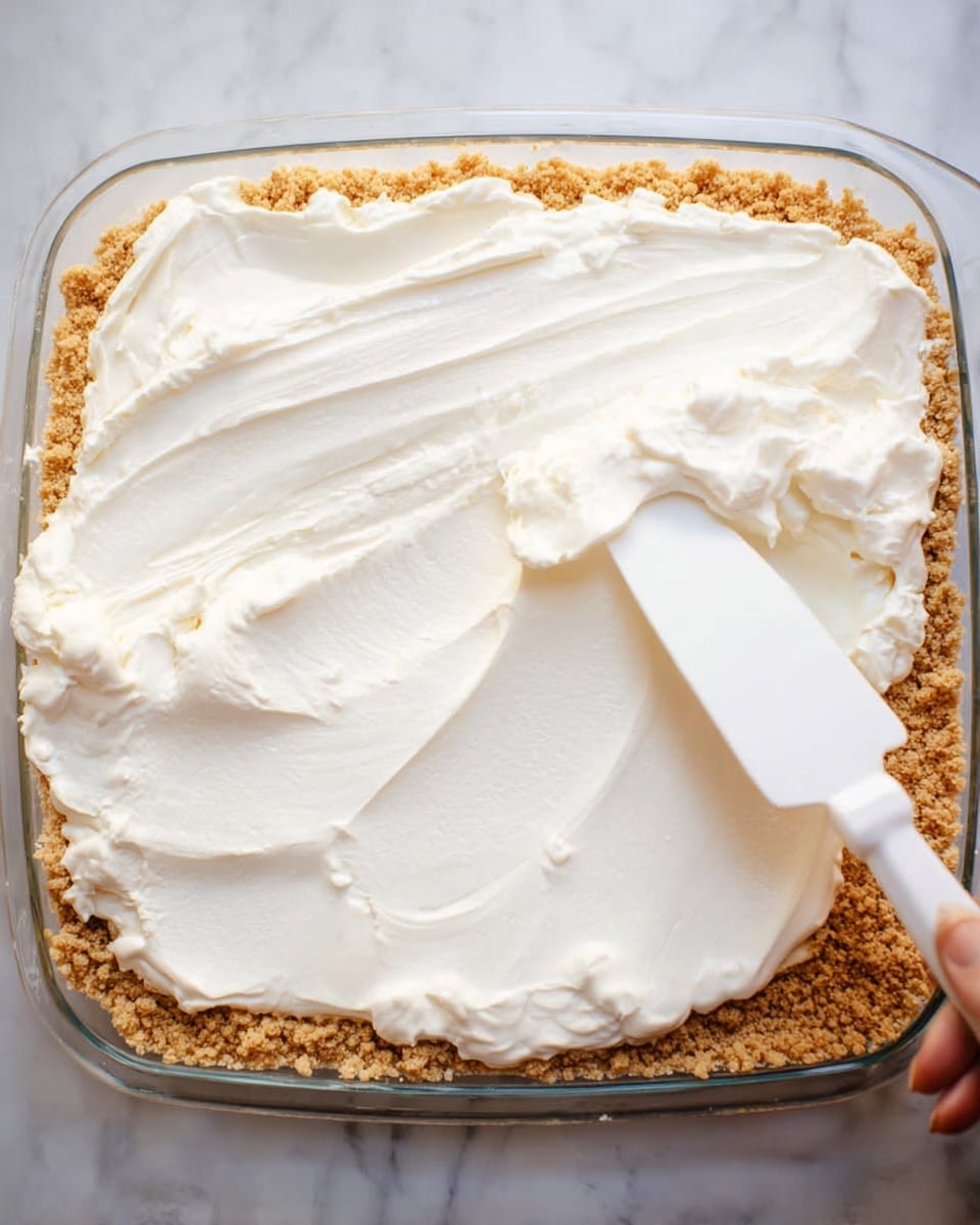 A clear glass dish contains two layers of a dessert: the bottom layer is a crumbly, light brown crust spread evenly, and the top layer is a thick, smooth white cream being spread with a white spatula held by a woman's hand, all placed on a white marbled surface. photo taken with an iphone --ar 4:5 --v 7