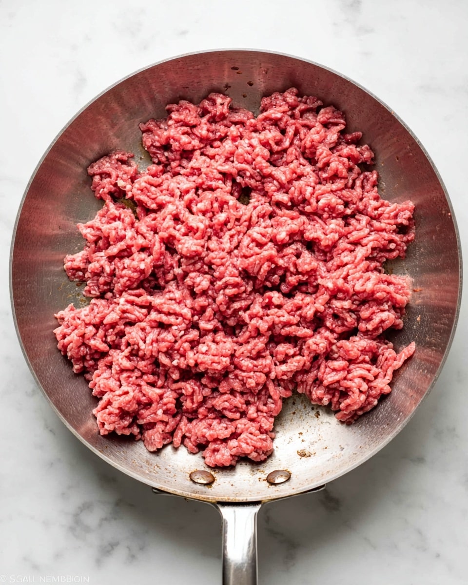 A round silver pan holds one layer of raw ground beef spread out mostly in the middle but with some pieces scattered unevenly around the edges, showing a bright red color with some parts turning light brown as it cooks. The pan's metal handle extends to the right, and the surface below is white marbled. The meat's texture looks crumbly and soft, with visible small chunks packed closely together. photo taken with an iphone --ar 4:5 --v 7