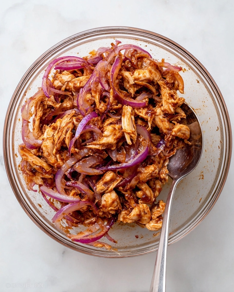 The image shows a clear glass bowl filled with a mix of shredded light brown chicken and thin slices of purple-red onion, all coated in a sticky, reddish-brown sauce. The chicken and onions are evenly mixed, giving the texture a combination of soft, juicy pieces with tender, crisp onion strips. A spoon with a metallic shine rests inside the bowl on the right side. The bowl sits on a white marbled surface. Photo taken with an iphone --ar 4:5 --v 7