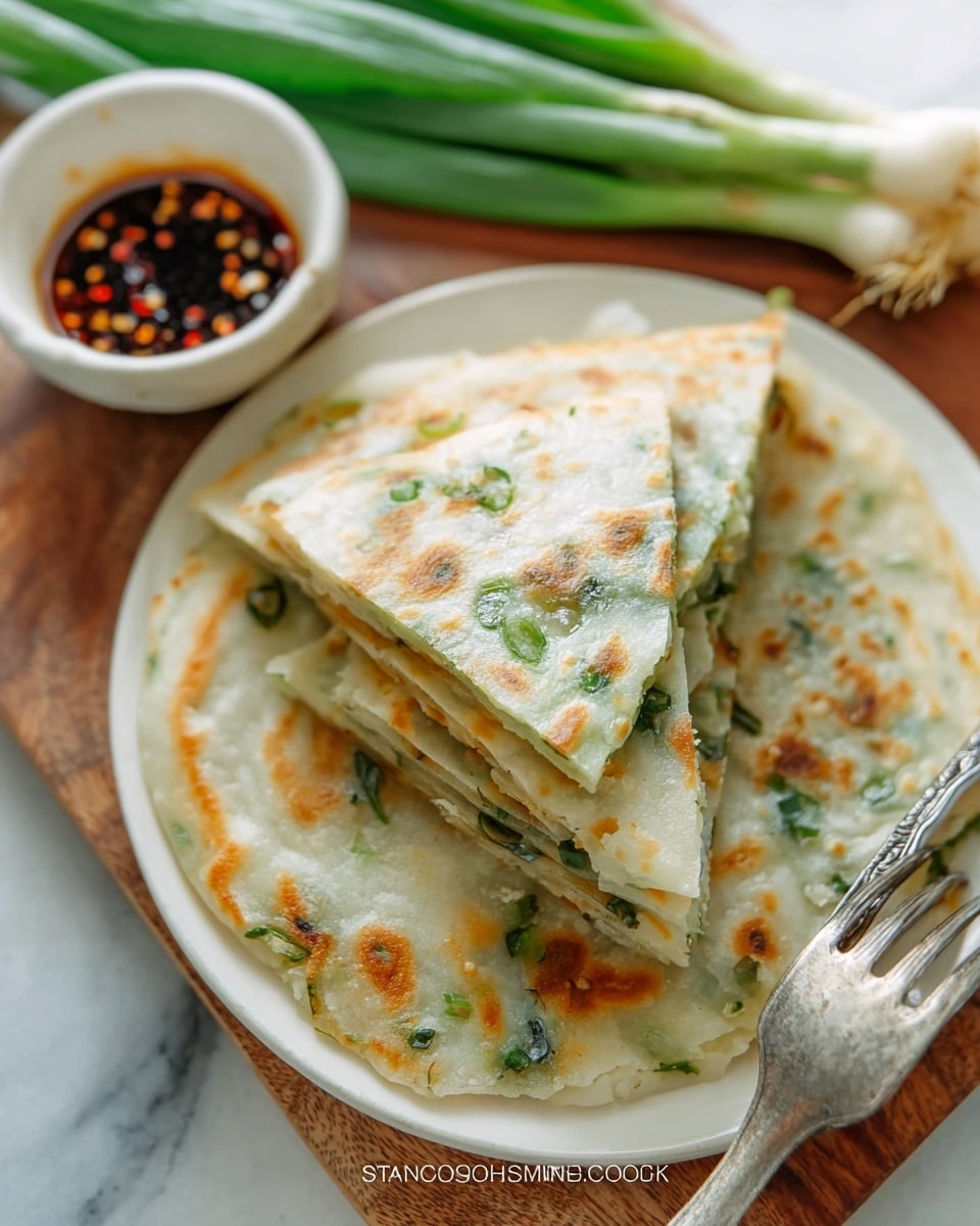The image shows layered green onion pancakes on a white plate, with three triangular pieces stacked on top of a whole round pancake. The pancakes have a light golden brown pattern from cooking, with small green bits of chopped onion visible inside the thin dough layers. To the side, there is a metal fork on the plate and a small bowl with dark dipping sauce sprinkled with chili flakes. Fresh green onions with roots still attached are placed nearby on a white marbled surface. photo taken with an iphone --ar 4:5 --v 7