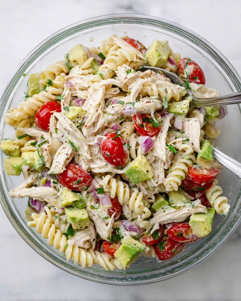 The image shows a clear glass bowl filled with a colorful pasta salad placed on a white marbled surface. The salad has three main layers: a base of creamy off-white spiral rotini pasta, chunks of light green avocado, and bright red cherry tomato halves mixed evenly throughout. Shredded pale chicken pieces with a slightly fibrous texture are layered on the salad, along with small bits of finely chopped red onion and green herbs sprinkled on top for freshness. The mixture looks well combined, with a few dark specks of seasoning visible, and two spoons are partially dipped in the bowl for serving. Photo taken with an iphone --ar 4:5 --v 7