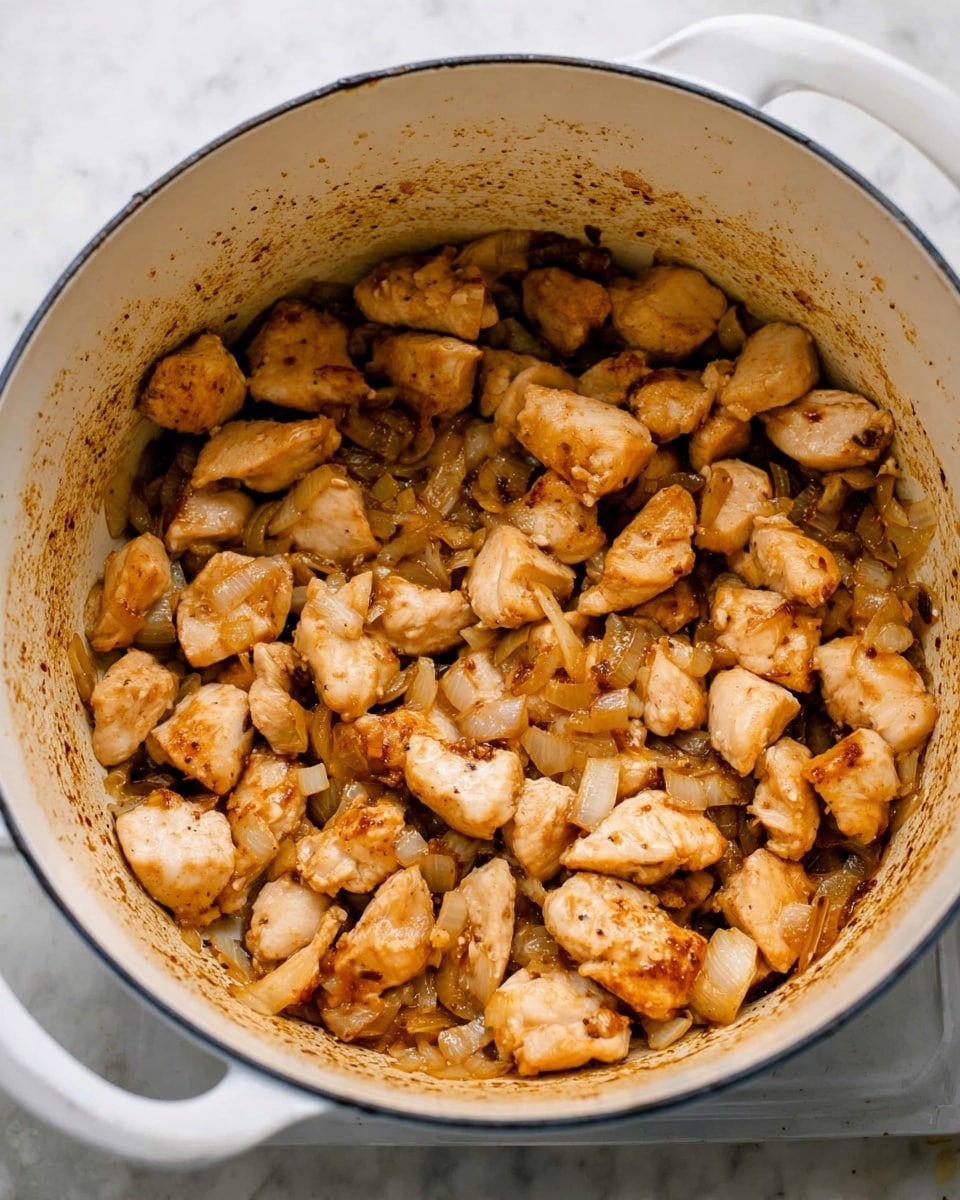 Inside a large white pot, many small pieces of light brown cooked chicken are mixed with translucent, slightly golden cooked onions. The chicken pieces have some browned edges giving a cooked texture. The sides of the pot show splashes of sauce residue in a darker brown color. The pot is seen from above, filled about halfway, sitting on a white marbled surface. photo taken with an iphone --ar 4:5 --v 7