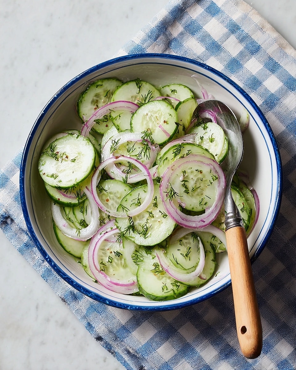 A white bowl with a blue rim holds a cucumber salad with thin round slices of pale green cucumber layered with thin curved slices of red onion that have a pale purple color. There are small bits of green dill sprinkled over the salad, adding a fine texture. A silver fork with a brown wooden handle rests inside the bowl on the right side, with cucumber slices on its tines. The bowl sits on a white marbled surface next to a white cloth with blue stripes. Photo taken with an iphone --ar 4:5 --v 7