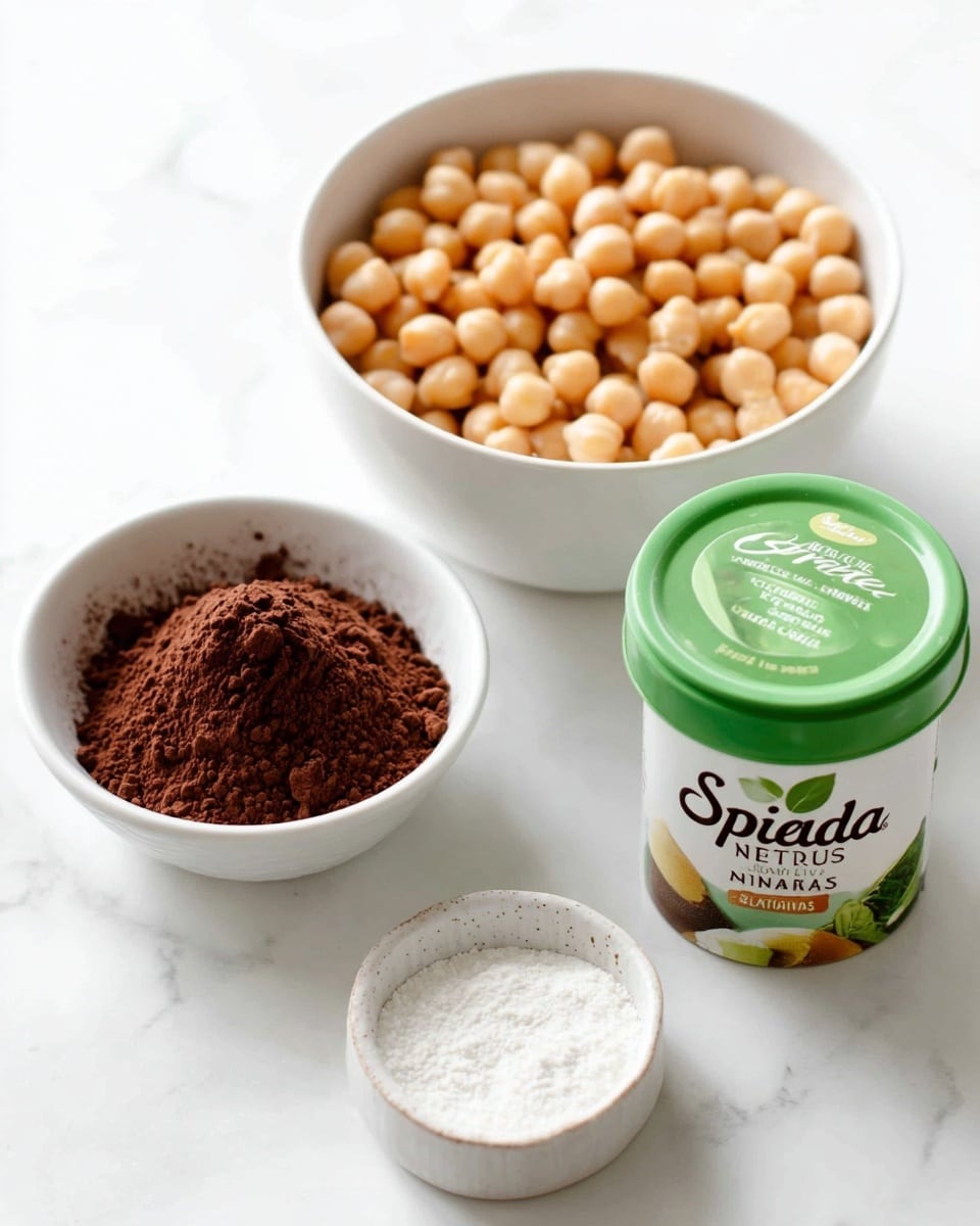 The image shows three white dishes and a container on a white marbled surface. In the back, there is a white bowl full of light tan round chickpeas. To its left, a smaller white bowl is filled with dark brown cocoa powder that looks soft and powdery. At the front right side, there is a small white dish with white granular powder. Next to it is a green-lidded container labeled