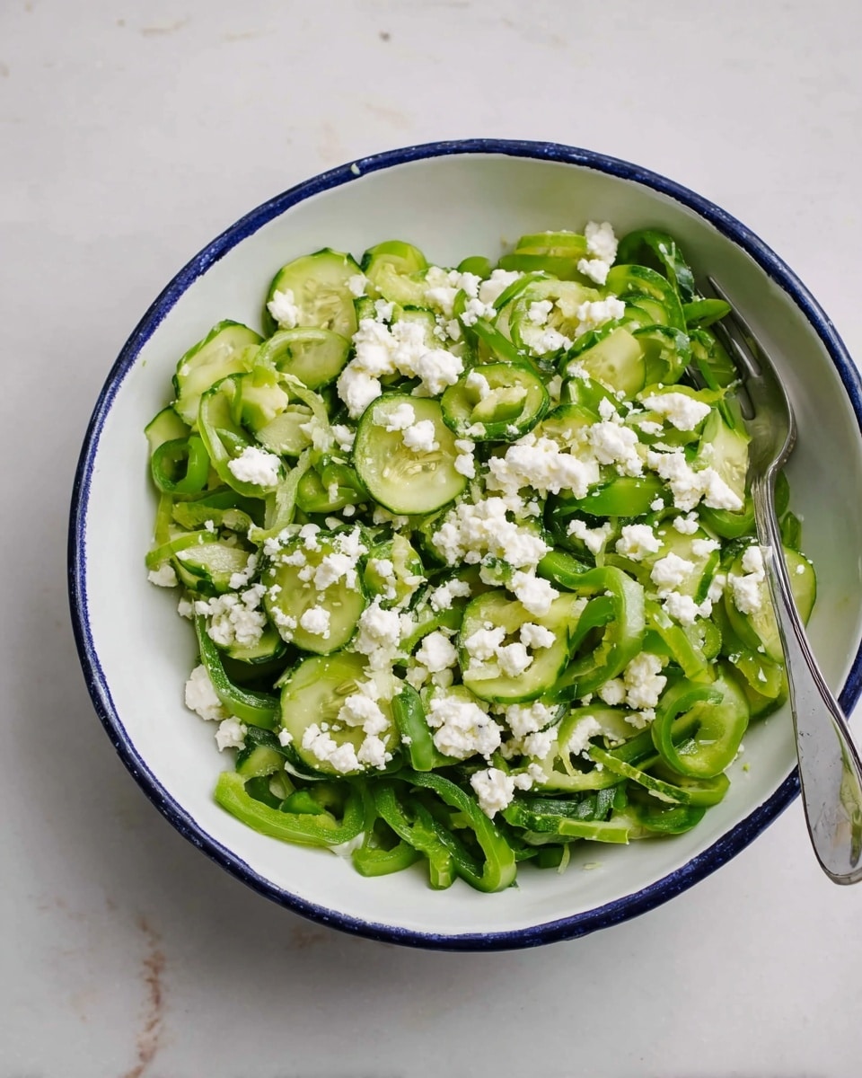 A white bowl with a thin blue rim holds a fresh salad on a white marbled surface. The salad has two main layers: the base layer consists of thinly sliced green cucumbers and thinly sliced green bell peppers, all mixed together. On top, there is a layer of white crumbled cheese scattered evenly over the vegetables. A silver fork rests inside the bowl on the right side. Photo taken with an iphone --ar 4:5 --v 7