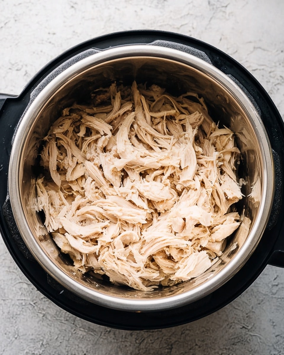 Inside a metal bowl of a black cooking pot sits shredded pale beige cooked meat. The meat pieces are layered loosely, with stringy and fibrous textures visible throughout. The bowl is shiny and clean, reflecting some light on the sides. The pot has a simple design with a black frame and buttons on one side. The background is a white marbled textured surface. photo taken with an iphone --ar 4:5 --v 7