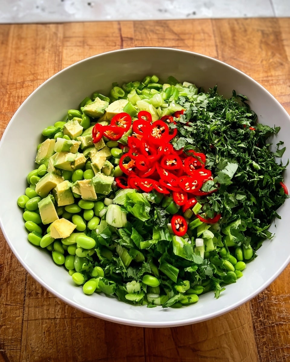 A large white bowl sits on a wooden surface with a white marbled texture edge showing, filled with several fresh layers of chopped green herbs and vegetables. The bottom layer is chopped dark green leaves, followed by chopped bright green spring onions. Above that, a large pile of light green edamame beans rests in the middle. To the left, chunks of green avocado add a creamy texture, and finely chopped dark green herbs fill the space on the right side. Thin slices of bright red chili peppers with visible seeds crown the center of the bowl, bringing a vibrant splash of color. The photo taken with an iphone --ar 4:5 --v 7